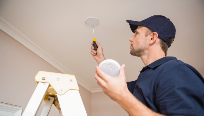 roofing contractor with dark blue shirt installing smoke detector with screwdriver on the ceiling